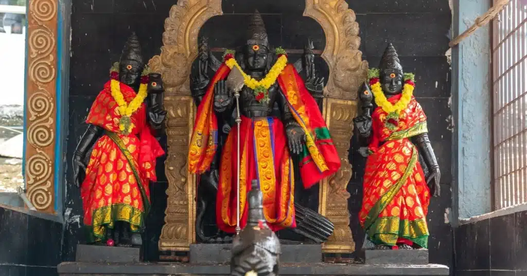 murugan history in tamil A temple scene featuring ornate black stone statues of deities, adorned with traditional attire and bright floral garlands.