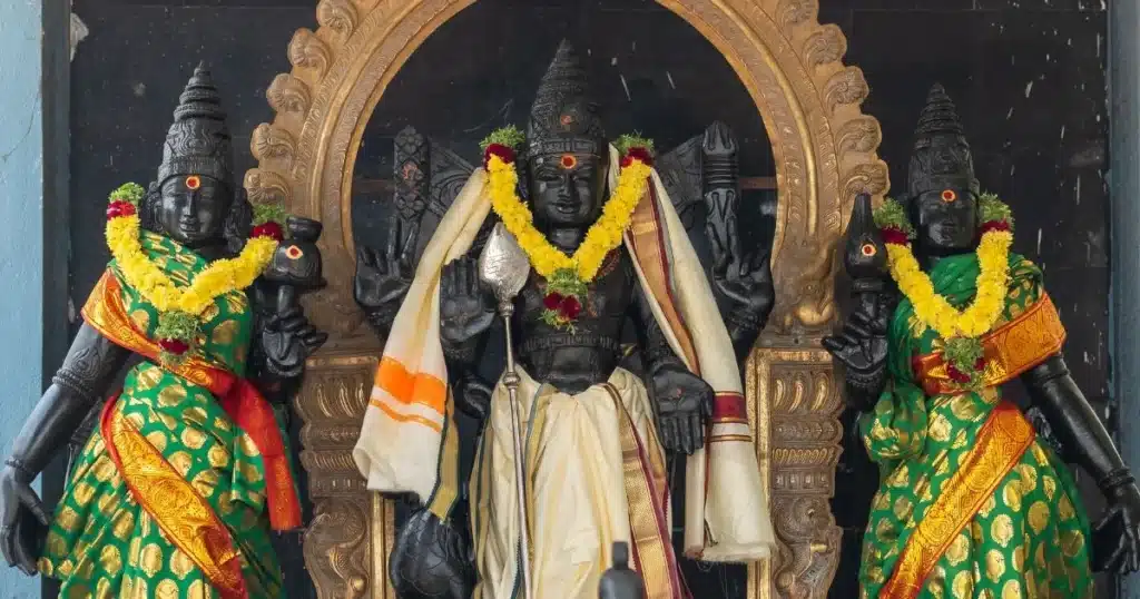 murugan history in tamil A striking temple scene featuring a black stone statue of Lord Venkateswara, flanked by two figures adorned in traditional attire and garlands.