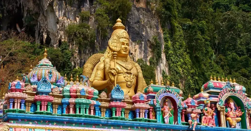 murugan history in tamil A large golden statue of Lord Murugan set against a natural backdrop of greenery and rock formations, with intricate temple architecture below.